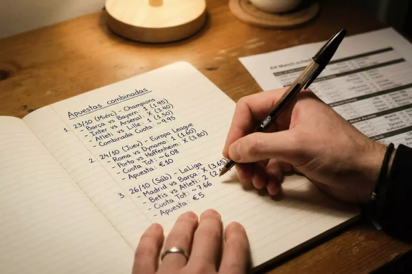 Mano sosteniendo un bolígrafo sobre una hoja con anotaciones de apuestas y estadísticas de fútbol