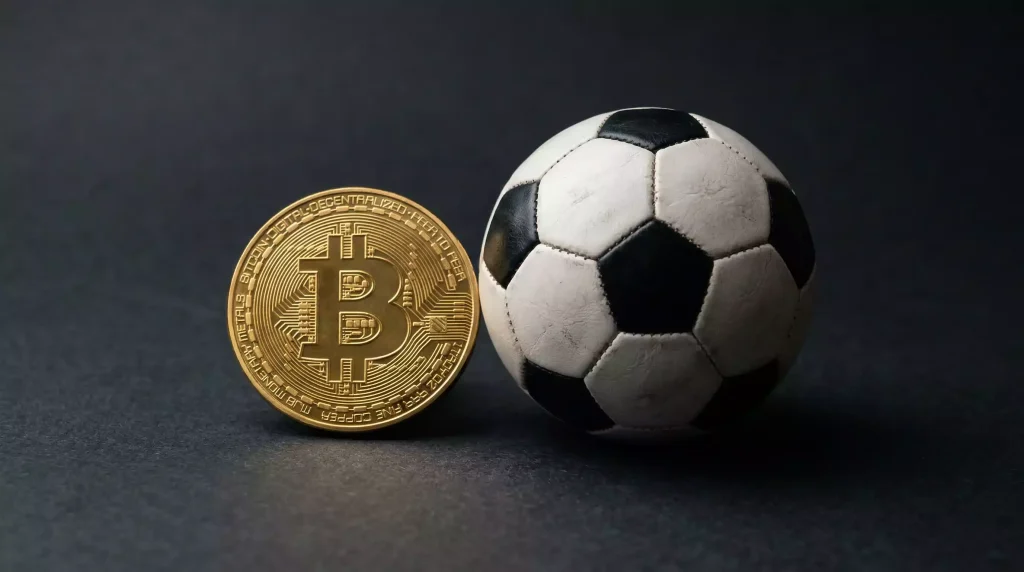 Moneda física de Bitcoin junto a un balón de fútbol sobre una superficie oscura