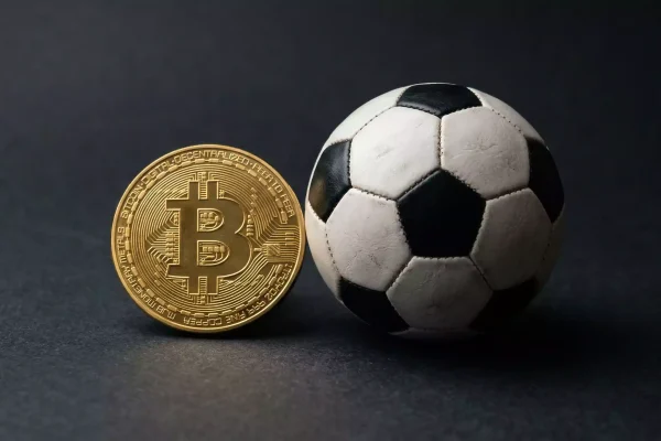 Moneda física de Bitcoin junto a un balón de fútbol sobre una superficie oscura