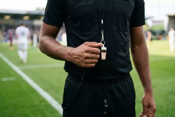 Árbitro de fútbol sosteniendo un silbato en un campo de juego con líneas de cal sobre césped verde